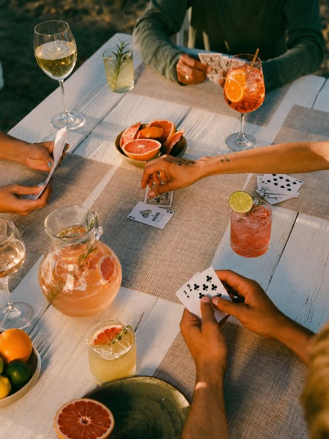 Friends playing cards around a sunlit table with spritzes and citrus