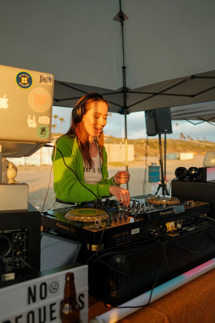 Marie DJing on the beach at golden hour, behind a sign that says no requests
